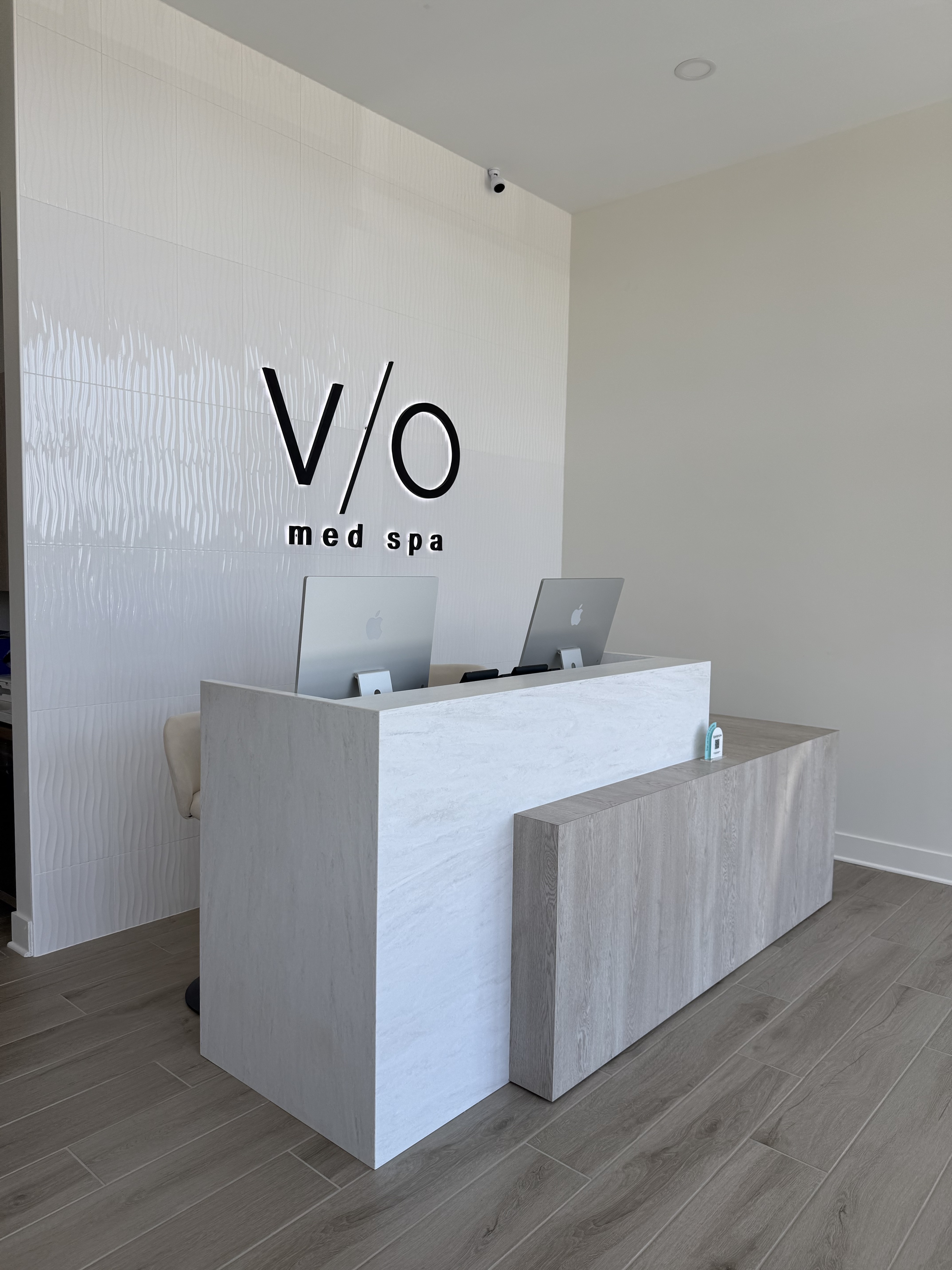 VIO Med Spa reception: V/O logo on a glossy textured feature tile wall, white marble reception desk with warm gray wood-grain accent panel, dual MacBook workstations, and warm gray plank flooring.